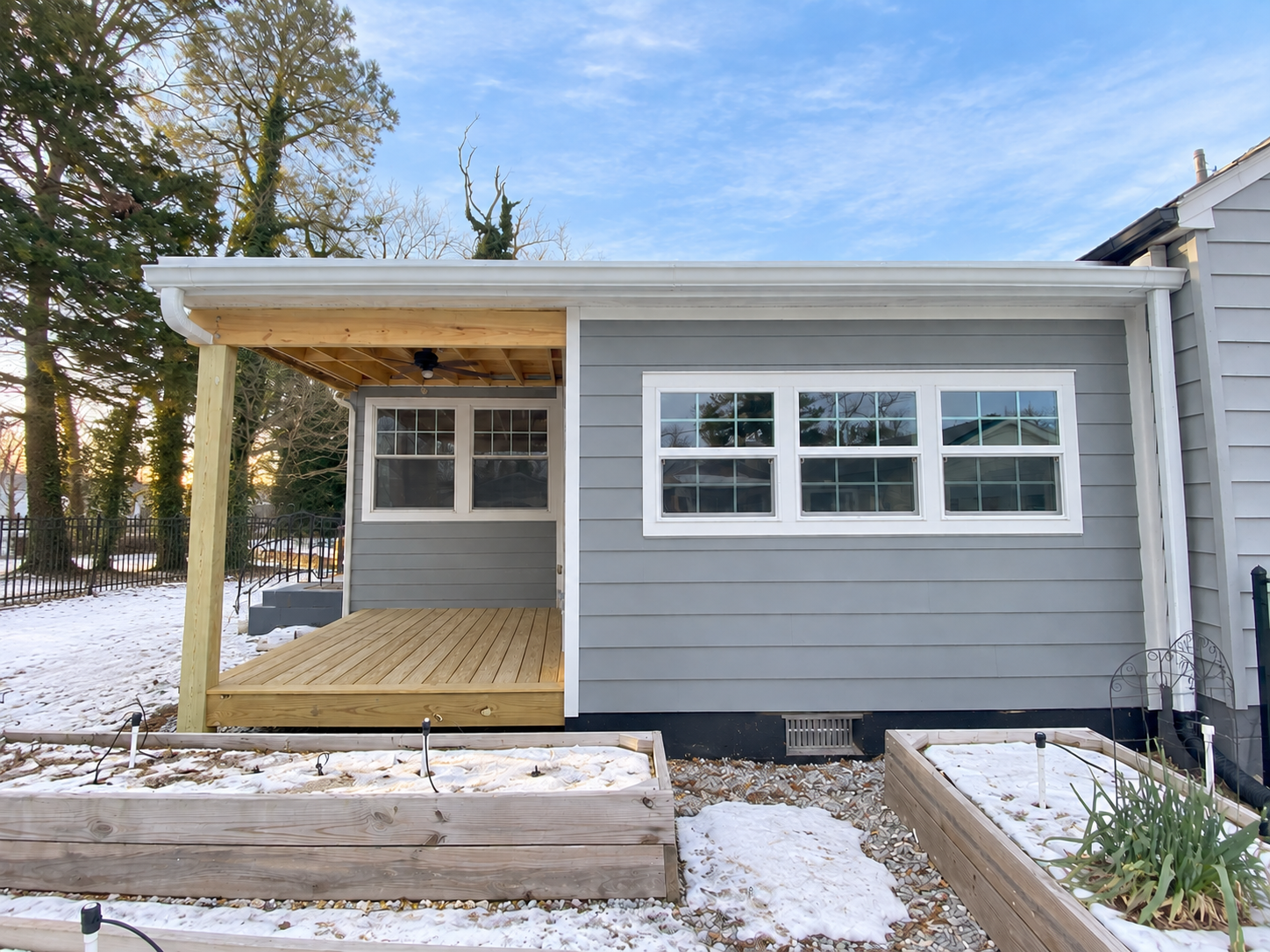 The back of a house with newly constructed wooden porch and windowed wall, surrounded by snow-covered garden beds and trees under a blue sky.