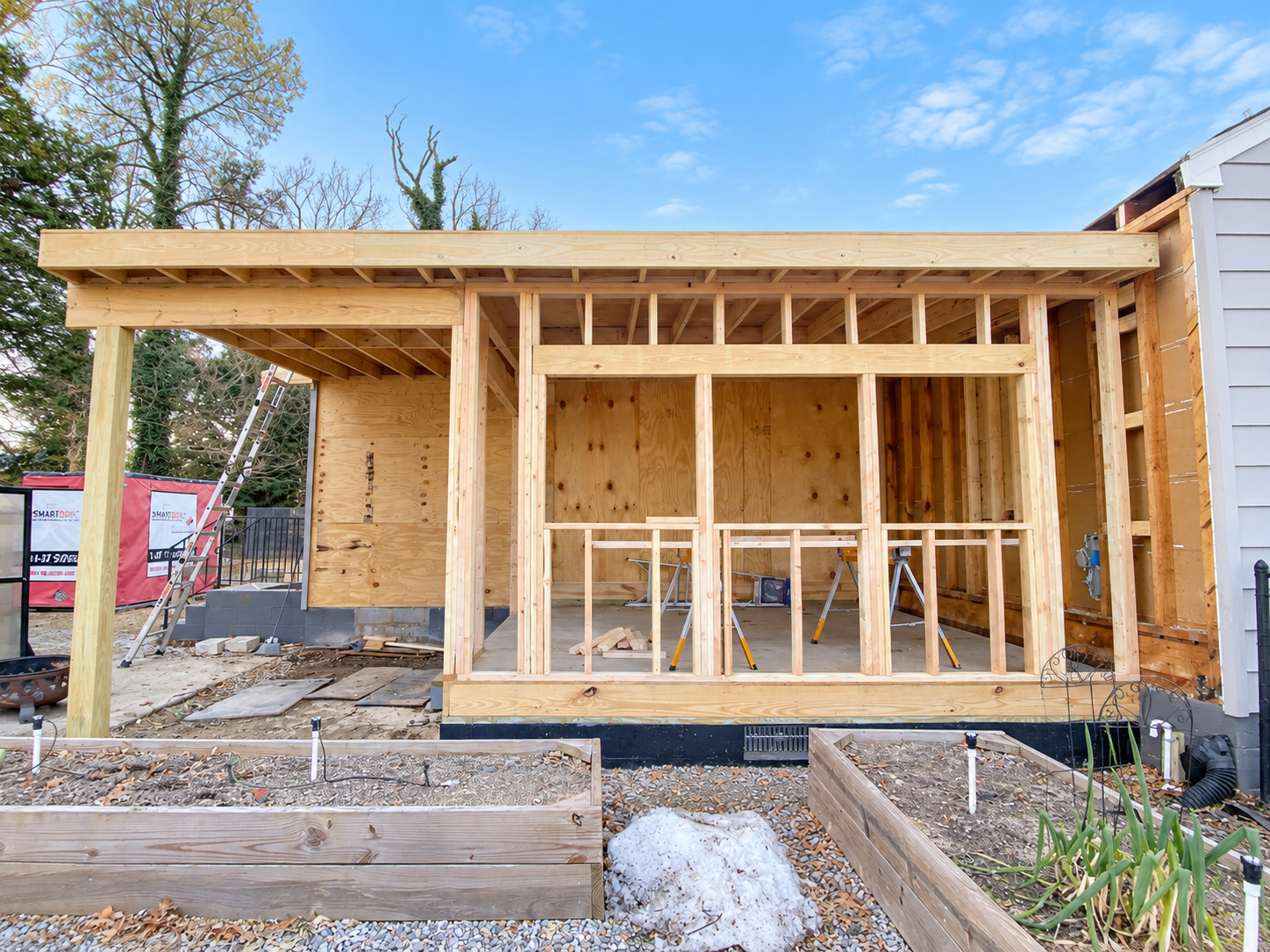 Wooden framework of a house under construction with some support beams, a ladder, and garden beds in the foreground.
