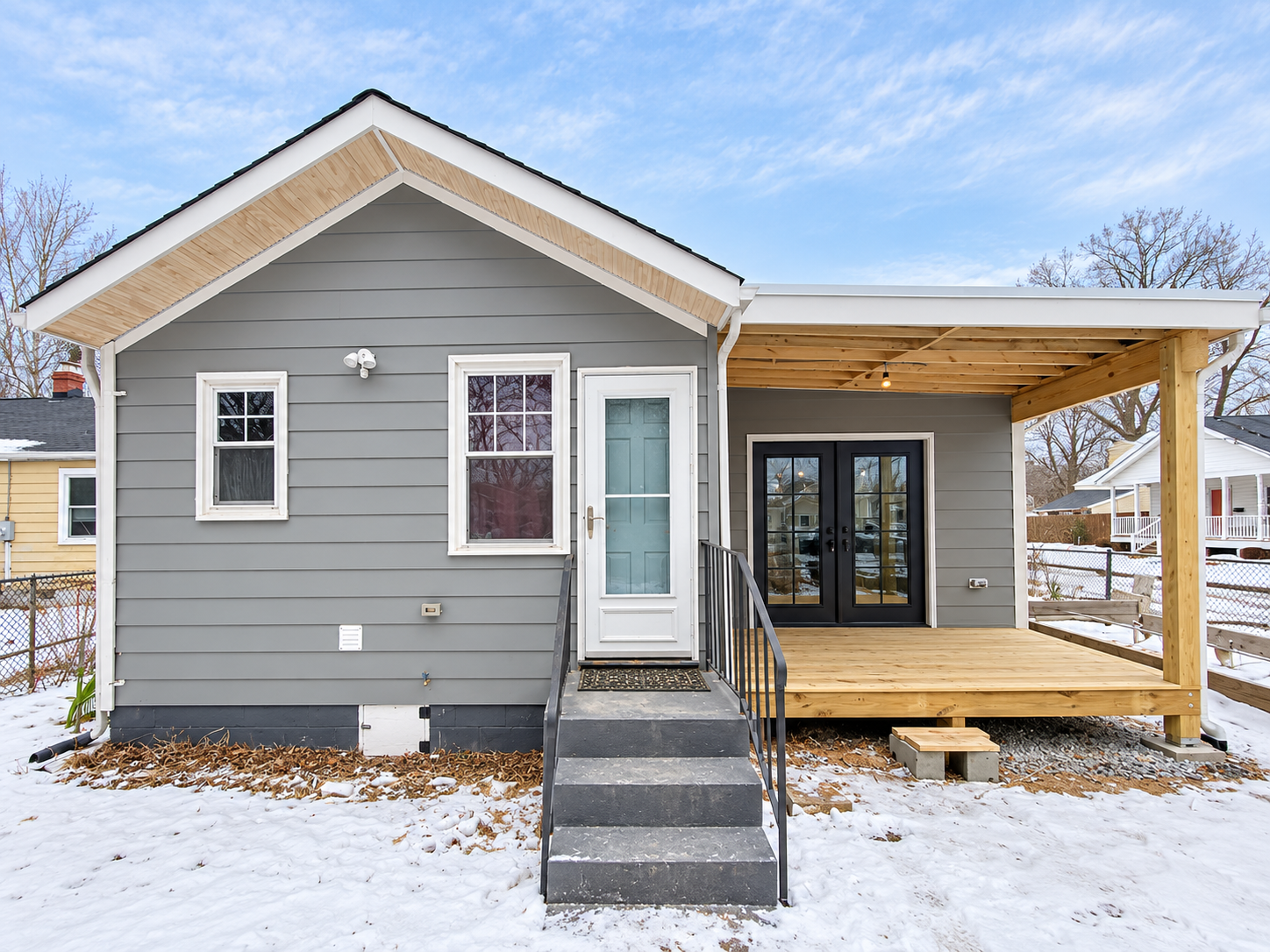 New gray house with white trim, small stairs leading to a front door, and a wooden deck under construction on a snowy day.