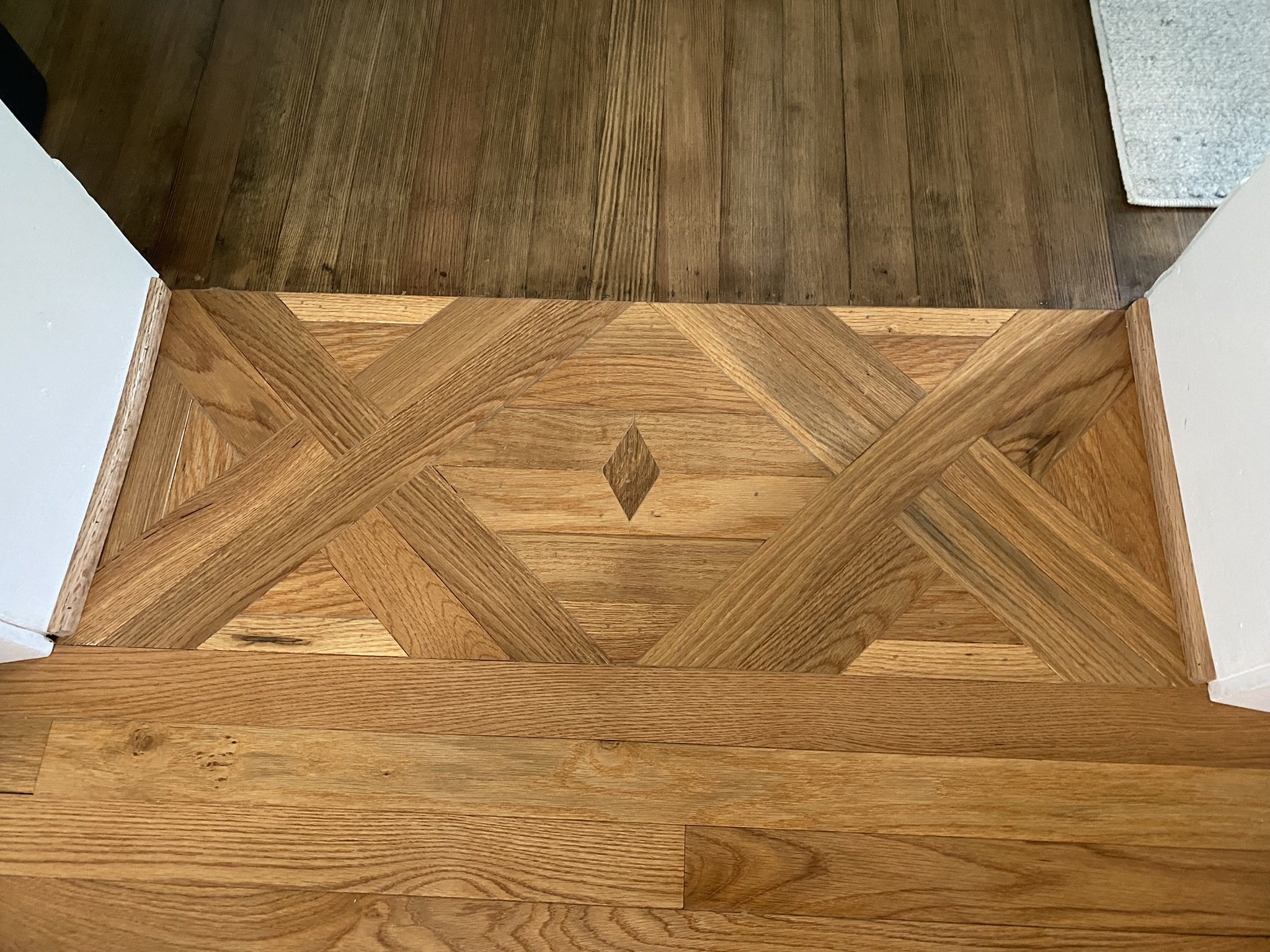 The image shows a corner of a room where hardwood flooring transitions from a parquet pattern to a straight plank pattern. There is a white wall on the left and right sides, and a beige area rug in the top right corner.