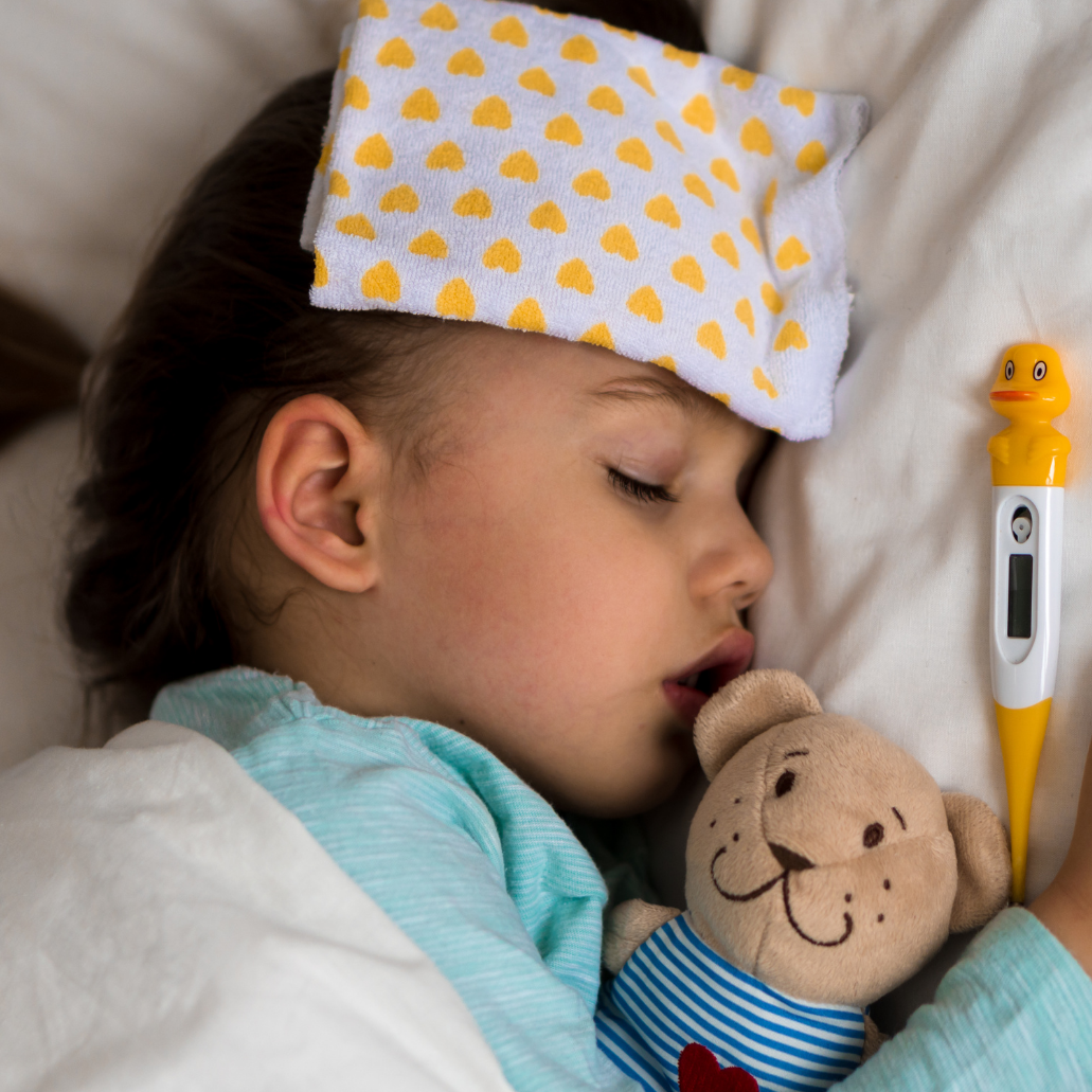 A young child sleeping in bed with a stuffed teddy bear, a rubber duck thermometer, and a cloth on their forehead.
