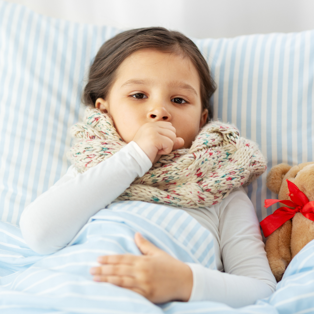A young girl lying in bed covered with a blue and white striped blanket, wearing a white long-sleeve shirt and a multicolored knit scarf, with a teddy bear beside her, resting her hand on her stomach and appearing to be unwell.