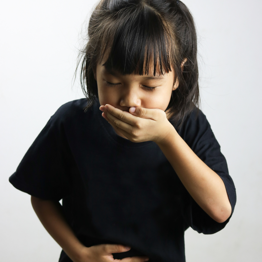 A young girl with dark hair and bangs, wearing a black shirt, appears to be feeling unwell, holding her stomach with one hand and covering her mouth with the other, against a plain white background.