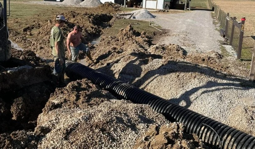 Workers installing a large black corrugated pipe in an excavated area of dirt and gravel.