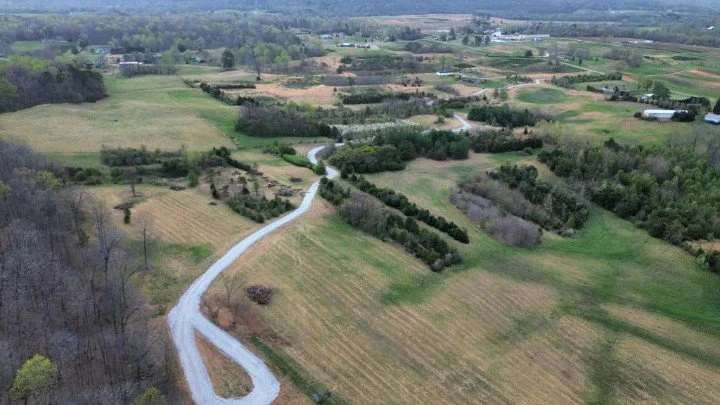 Aerial view of a rural landscape with a winding gravel road, open fields, patches of trees, and scattered farm buildings in the distance.