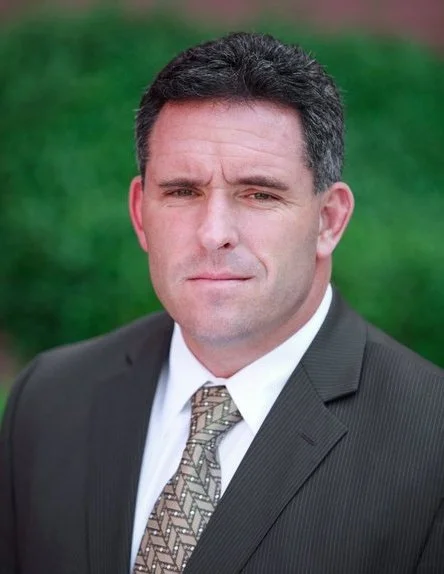 Portrait of a man in a dark suit and patterned tie standing outdoors with a blurred green background.