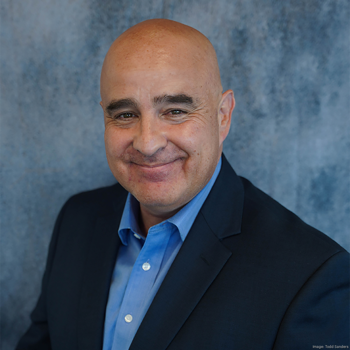 Headshot of a smiling bald man in a blue shirt and dark blazer, against a blue textured background.