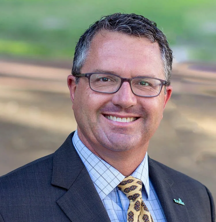 A middle-aged man with glasses, wearing a dark suit, light blue shirt, and a patterned tie, smiling outdoors with a blurred natural background.