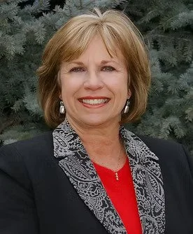 A smiling woman with short light brown hair, wearing a dark blazer, a red top, and a patterned scarf, standing outdoors with green foliage in the background.