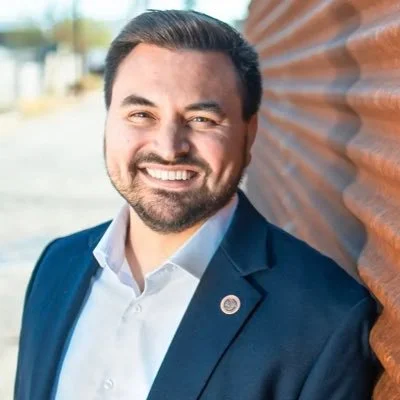 A man with dark hair and a beard smiling, wearing a blue suit jacket and white shirt, standing outdoors next to a rust-colored corrugated metal wall.