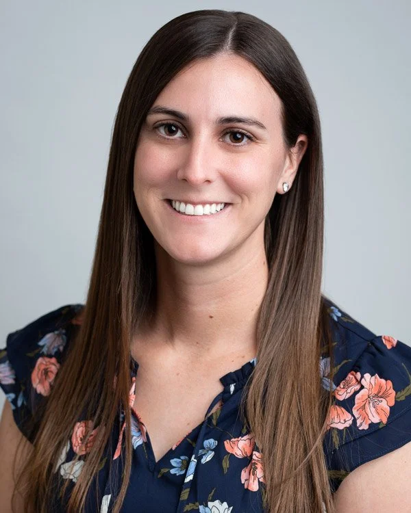 Portrait of a young woman with long brown hair, smiling, wearing a navy floral blouse and small pearl earrings, against a plain light gray background.