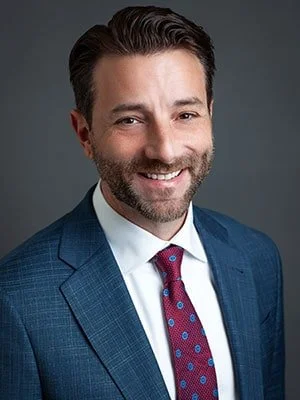A professional man with dark hair and a beard, wearing a blue suit and a red patterned tie, smiling against a gray background.