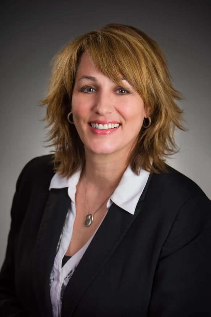 Professional headshot of a woman with shoulder-length reddish-brown hair, smiling, wearing a black blazer and white blouse, with a necklace and earrings, against a gray gradient background.