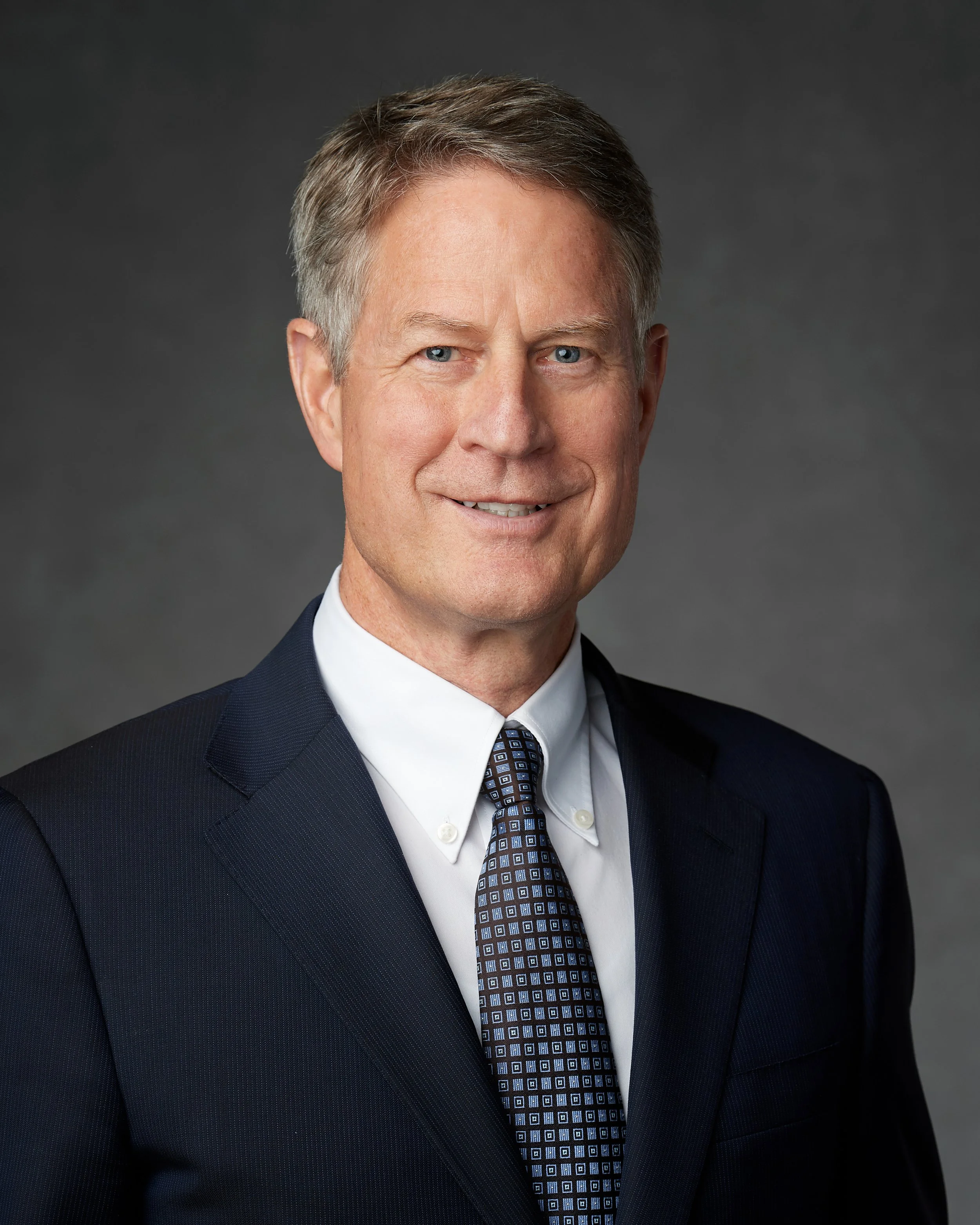 A professional man with gray hair wearing a dark suit, white shirt, and patterned tie smiling against a gray background.
