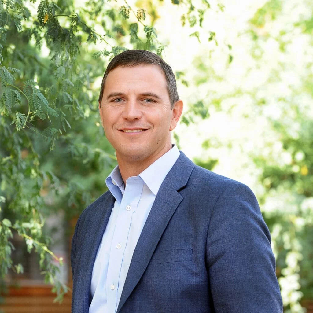 A man wearing a blue blazer and white dress shirt, smiling outdoors with greenery in the background.