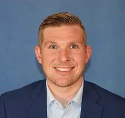 Portrait of a young man with short blonde hair, wearing a navy blazer and light blue shirt, smiling against a blue background.