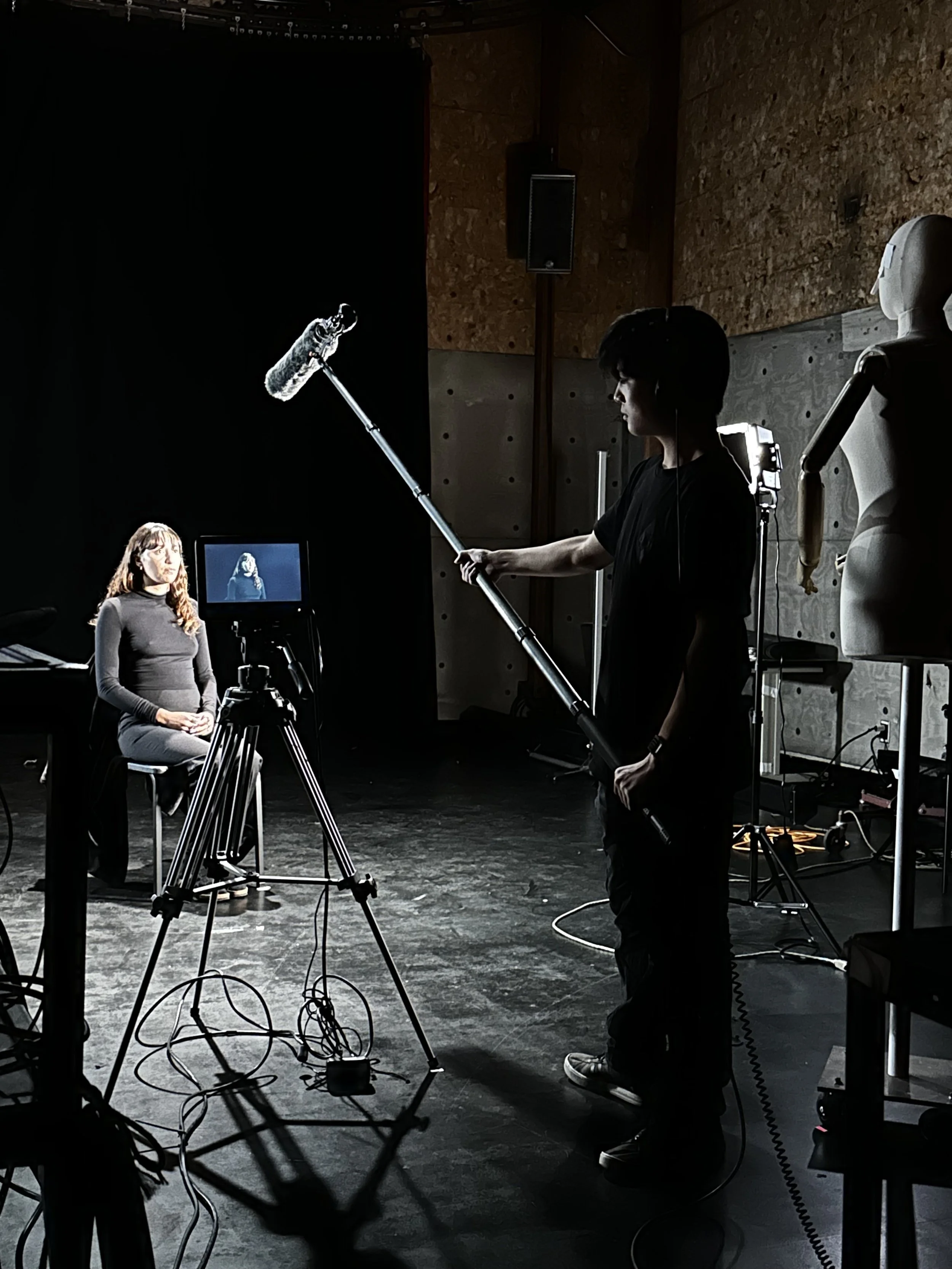 Filmmaker filming a woman in a recording studio with a boom microphone and video camera.