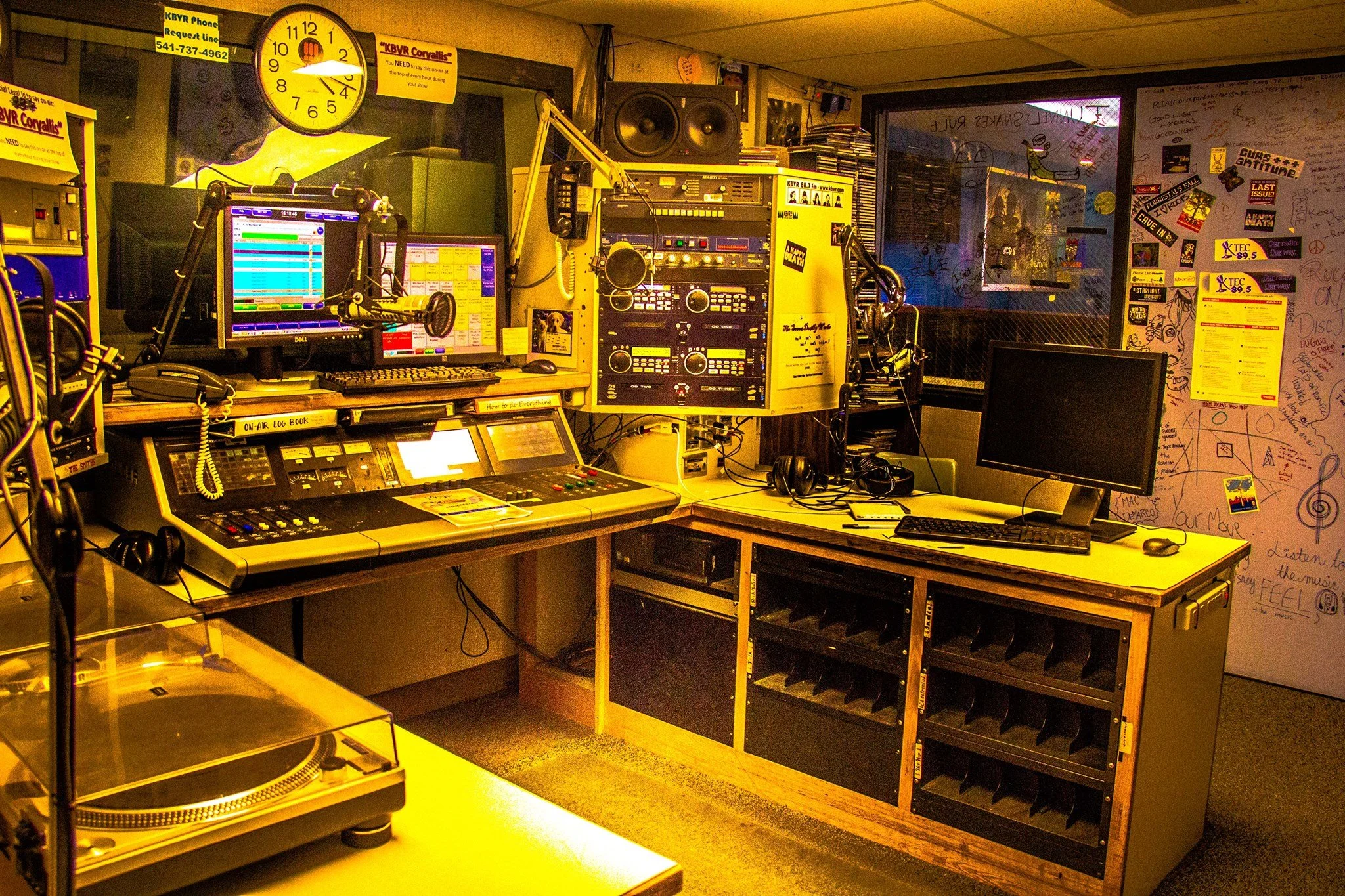 radio recording studo, with KBVR corvallis stickers and overally saturated, yellow, contrast photo edit. Large clock and rack mounted audio visual equiptment abound, Aqua Ideation LLC WCAG prompt reviewed.