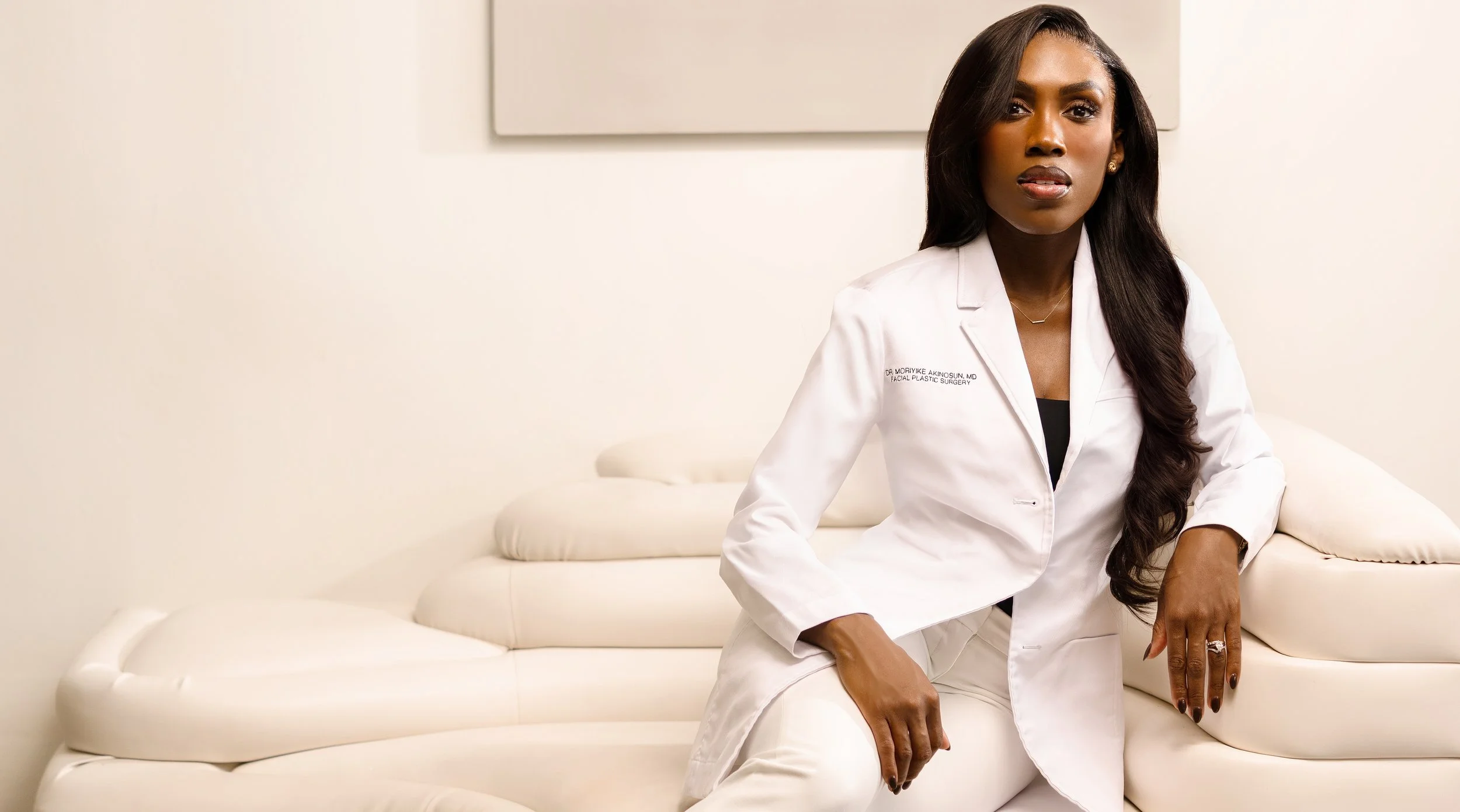A professional woman with long dark hair, wearing a white lab coat, sitting on a white leather couch in a minimally decorated room.