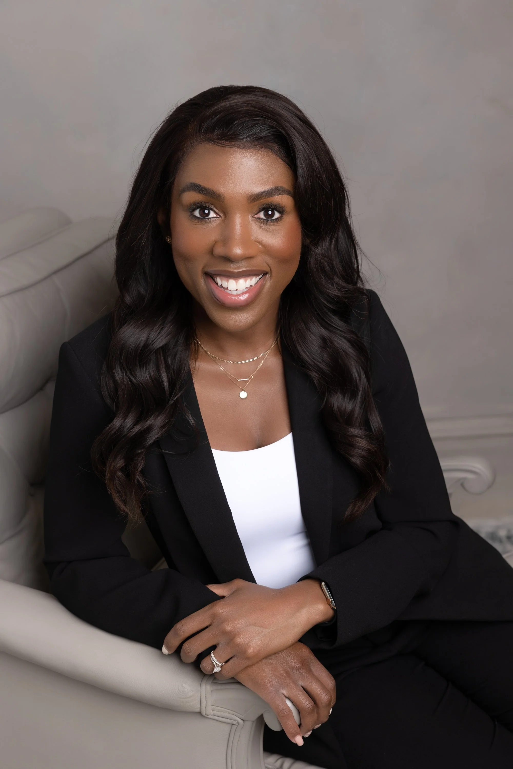 A woman with long dark wavy hair, wearing a black blazer and white top, smiling, sitting on a light-colored sofa, with jewelry and a watch, in a professional portrait setting.
