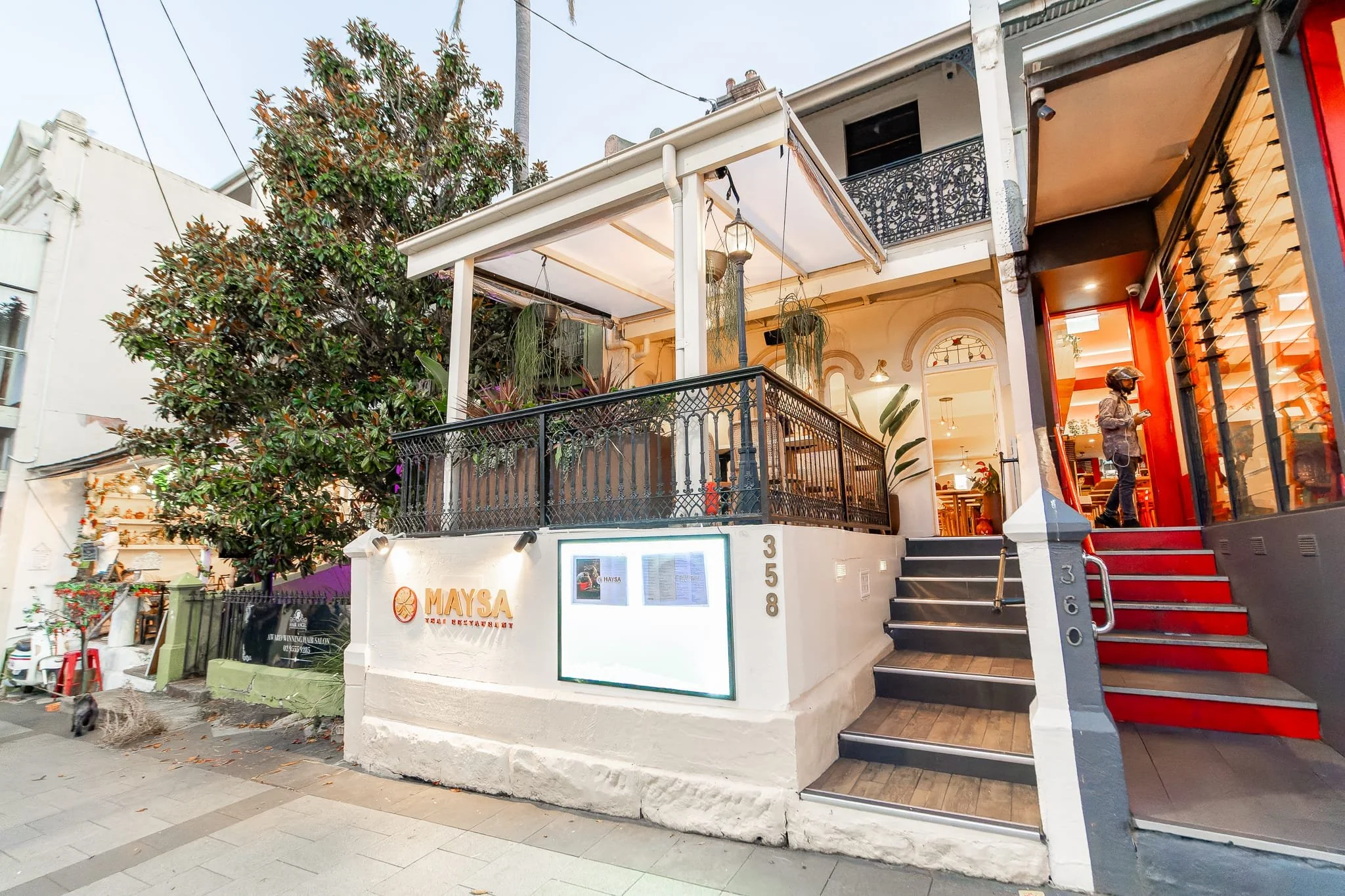 The exterior of a two-story restaurant with a white facade, black metal balcony, and green plants on the balcony porch. The restaurant's name "MAYSA" is displayed on the front wall, with the address 358 nearby. Red stairs lead up to large glass doors, through which a person is visible inside. Street and sidewalk with nearby trees and buildings are also visible.