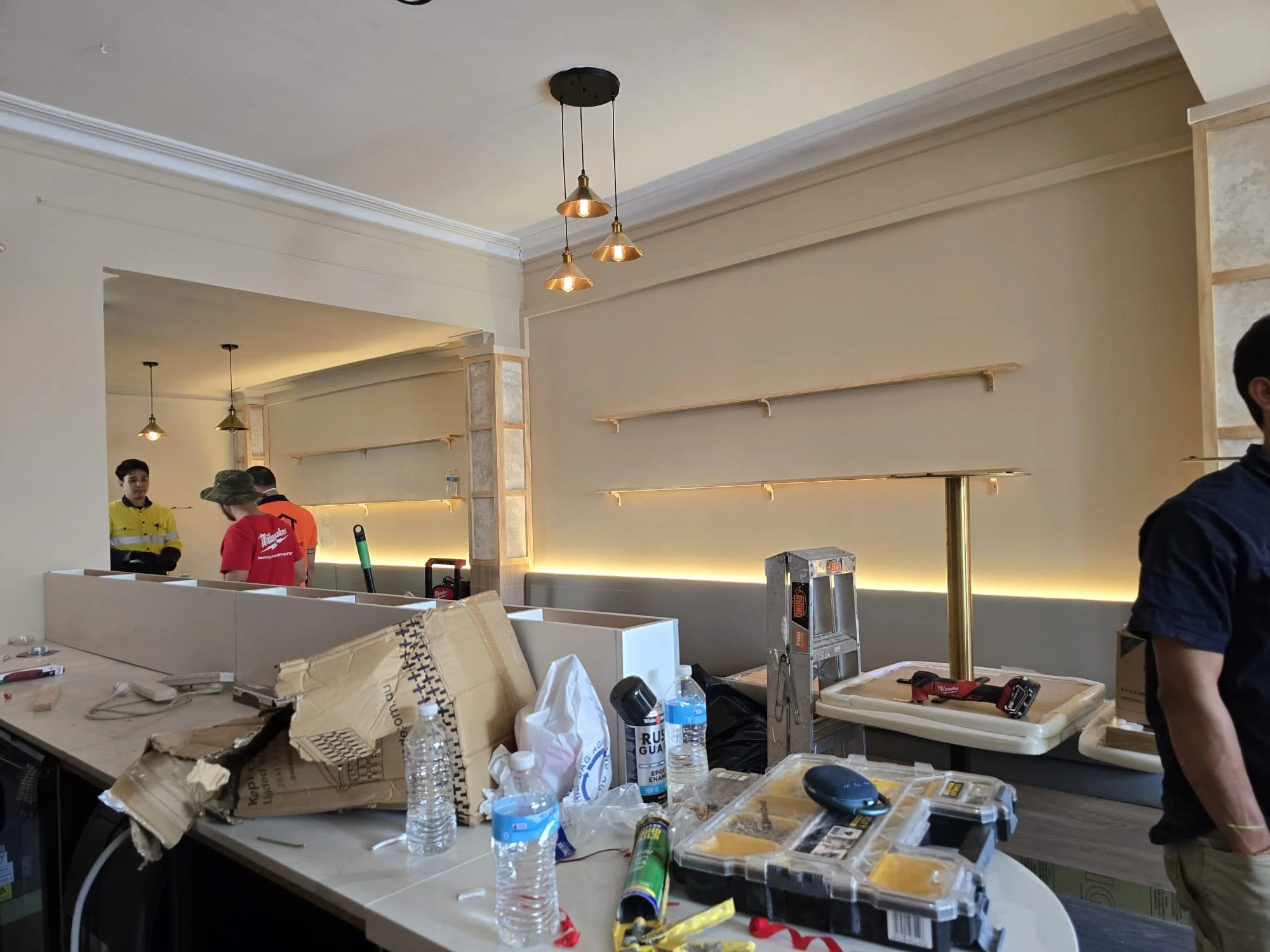 Interior of a restaurant or café under construction with cluttered countertop, water bottles, tools, and workers in the background.