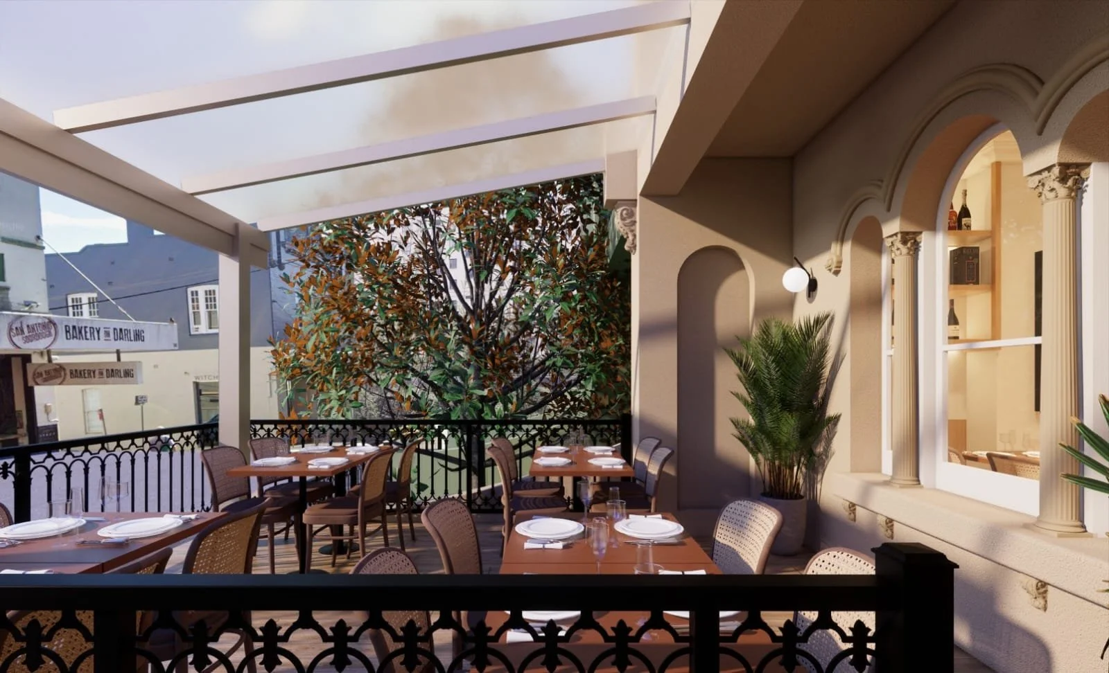 Outdoor restaurant patio with tables set with white plates, glasses, and utensils, surrounded by a decorative black fence, with a large potted plant and trees in the background, and an arched window with columns on the side wall.