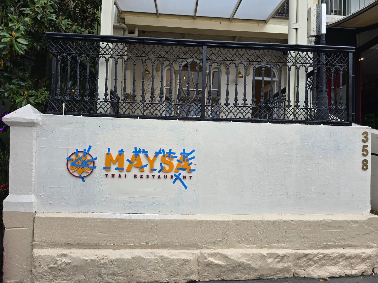 The exterior wall of a Thai restaurant named MYASE, with the address 358, featuring a black metal railing balcony above and a colorful sign with Thai motifs.