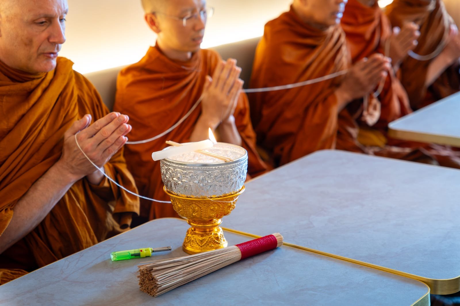 Four Buddhist monks in prayer, with hands pressed together, sitting at a table with a ceremonial object that has a lit candle on top, incense sticks, and a long, thin stick with a red grip.