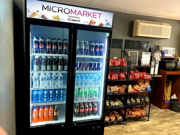 A vending refrigerator with bottled drinks and water in front of a snack shelf stocked with chips and snacks in a convenience store. A sign above reads 'MICRO MARKET powered by DIGIT'.