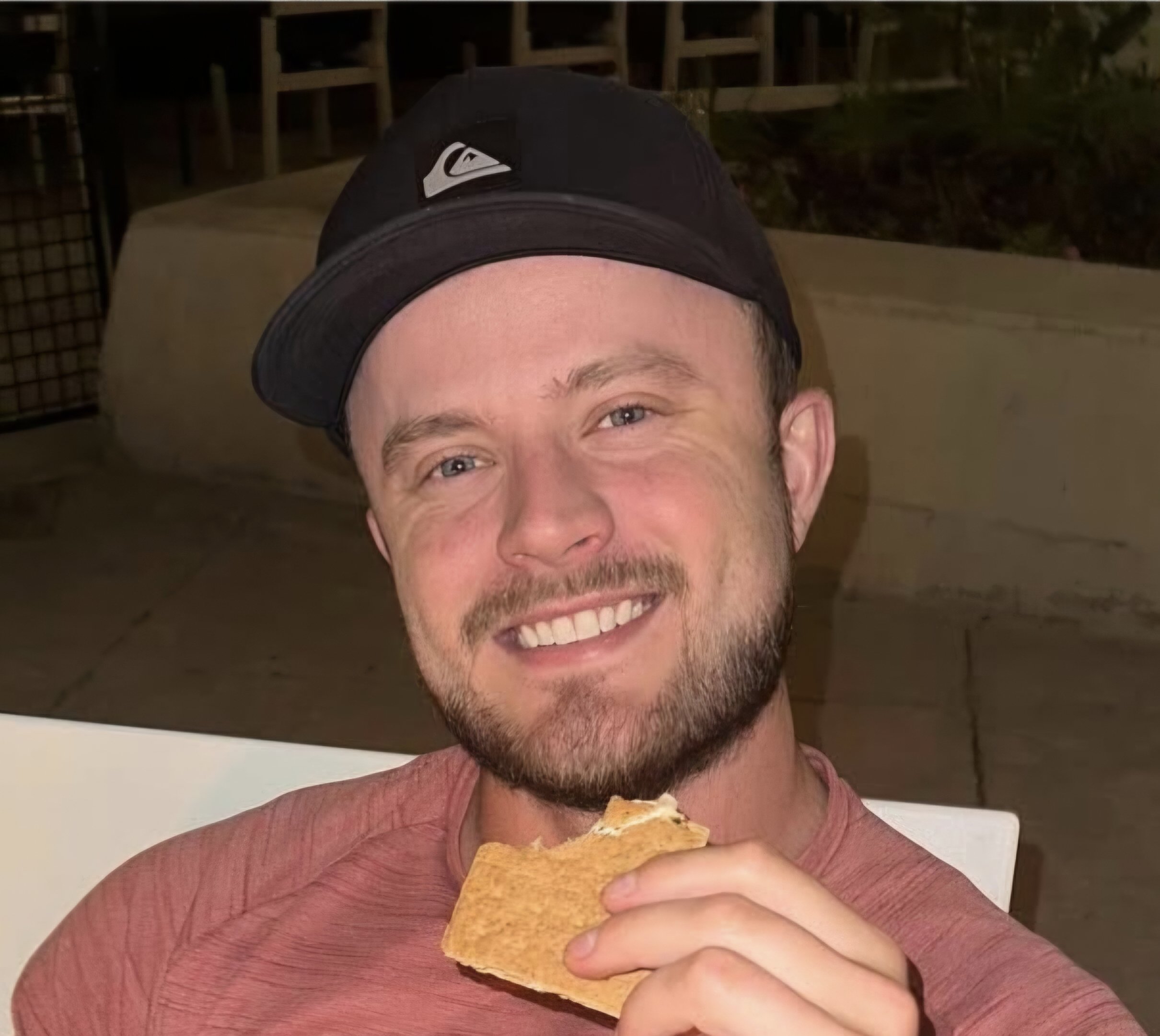 A smiling man wearing a black cap and a pinkish t-shirt, holding a taco near his mouth in an outdoor setting at night in Salt Lake City, Utah.