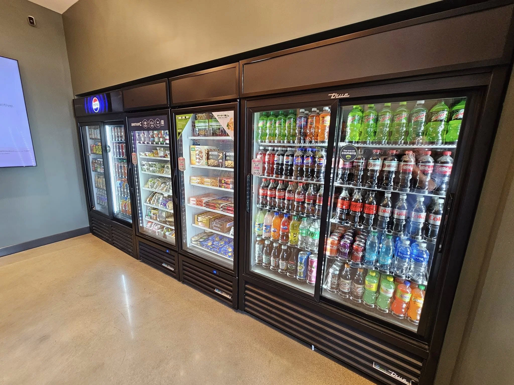 A row of refrigerated vending machines filled with various beverages and snacks in a retail or convenience store. The rightmost section contains bottles of soda like Coca-Cola, Sprite, and Fanta, while the left sections hold snacks and other items.