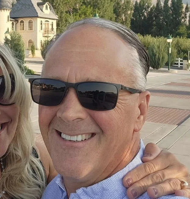 Close-up of a smiling middle-aged man wearing sunglasses and a blue collared shirt outdoors during daytime with trees and a building in the background in Salt Lake City, Utah.
