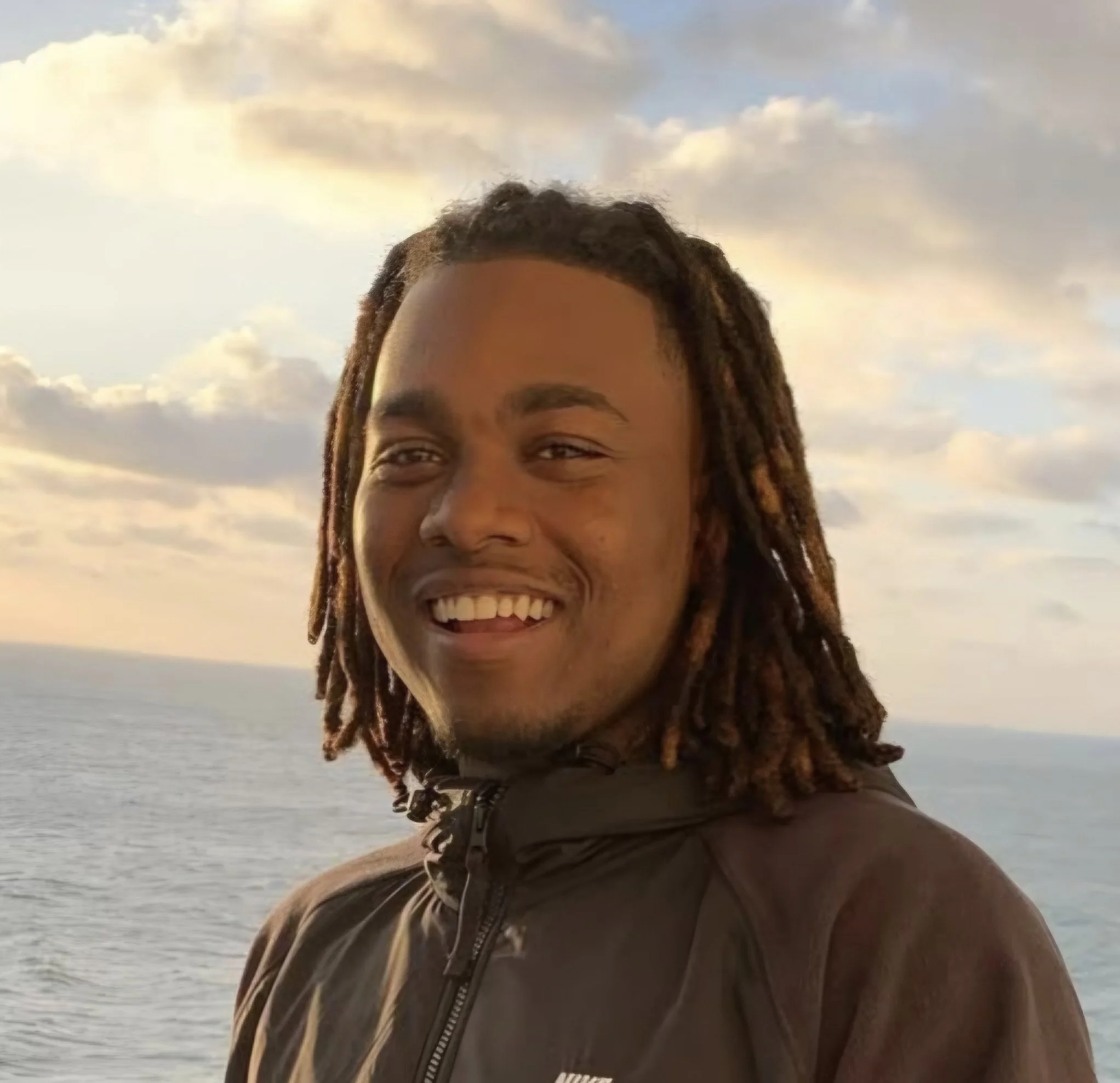 A smiling man with dreadlocks wearing a dark jacket stands outdoors near the ocean during sunset with clouds in the sky In Salt Lake City, Utah.
