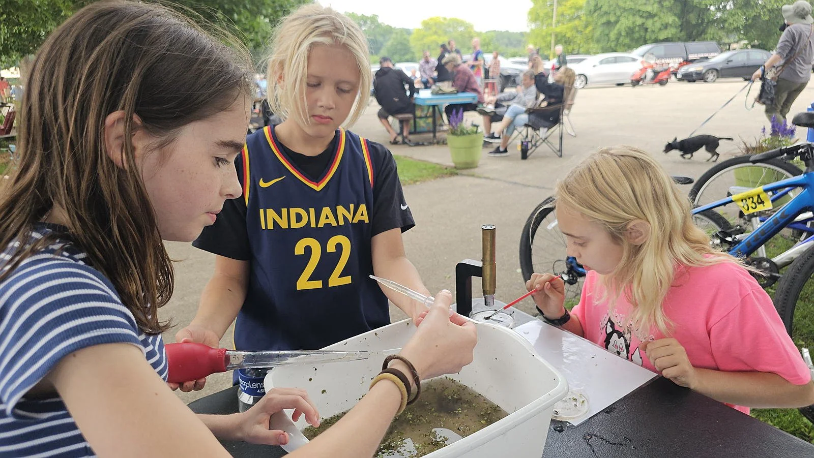 Three girls are conducting a science experiment outdoors at a table, with one holding a dropper and another using a tool near a small container. In the background, there are people sitting and chatting at picnic tables, with parked cars and a person walking a dog nearby.