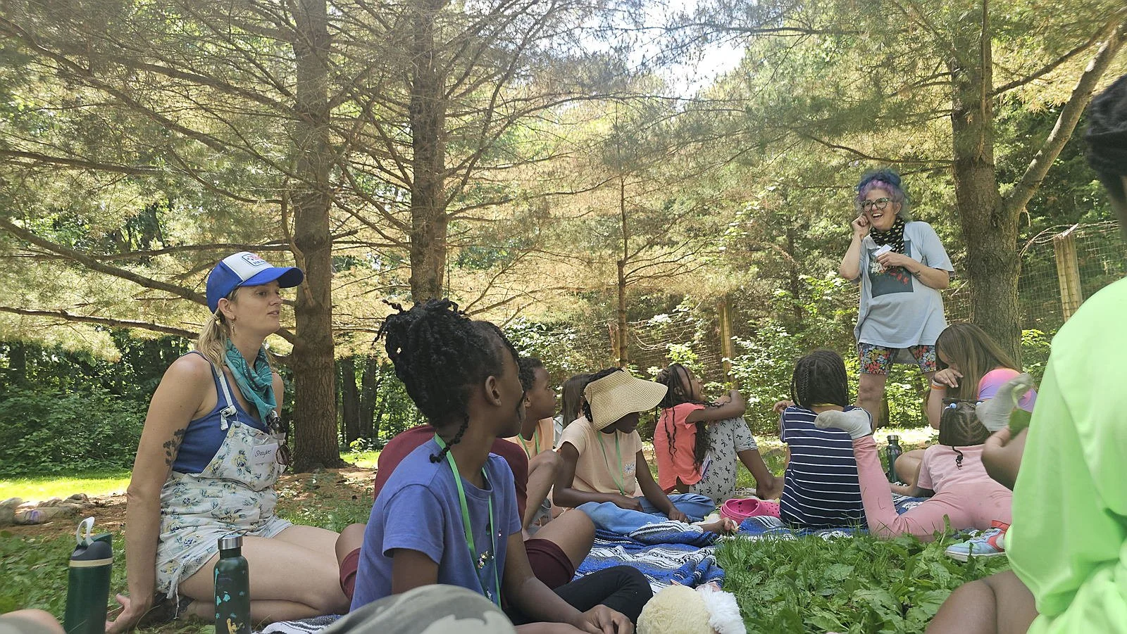 Group of children and adults sitting on the grass during an outdoor activity in a wooded area. One woman with purple hair and glasses is speaking or leading the group, while the others sit on blankets or directly on the grass, listening attentively."