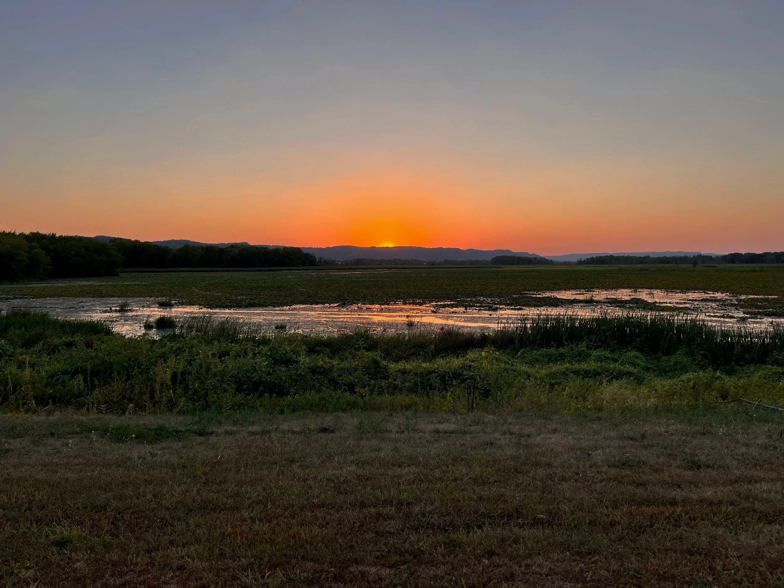 Sun setting over a river with surrounding greenery and hills in the distance.