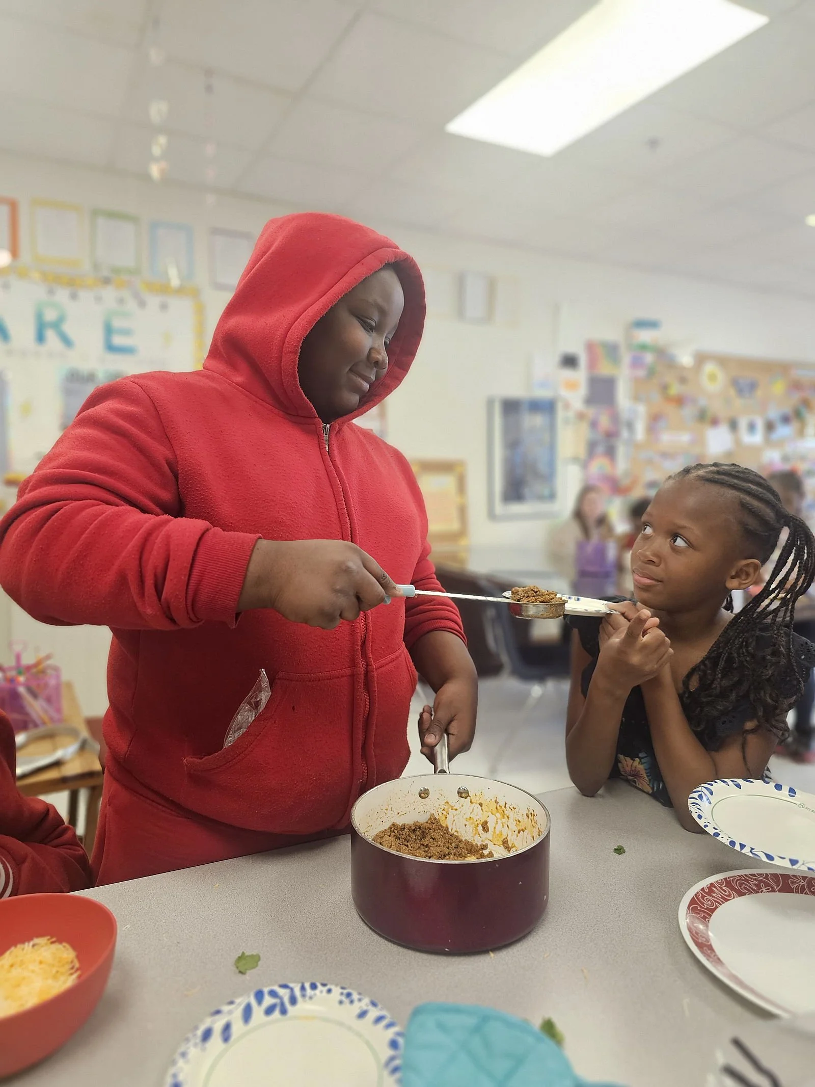A young girl with braided hair looks at a woman in a red hoodie serving food at a table. The woman is scooping food from a pot, and there are plates and bowls on the table. The background shows a decorated classroom with children.