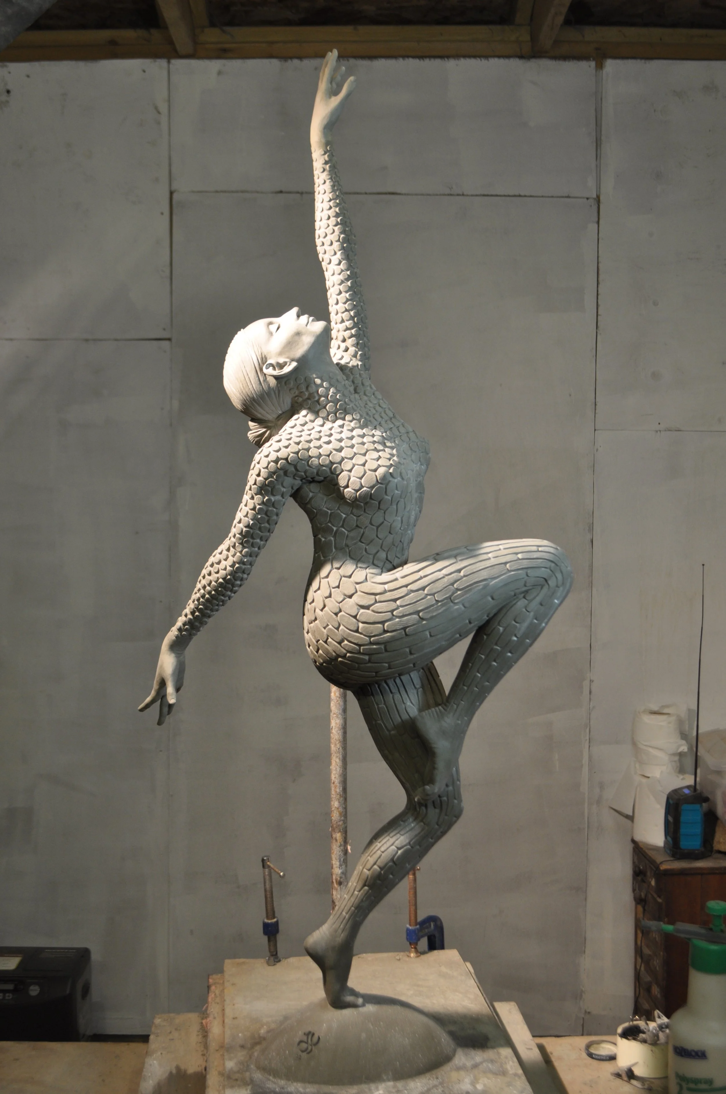 A sculpture of a woman with a honeycomb textured body, in a graceful pose with one arm raised and head tilted back, mounted on a base in an artist's studio.
