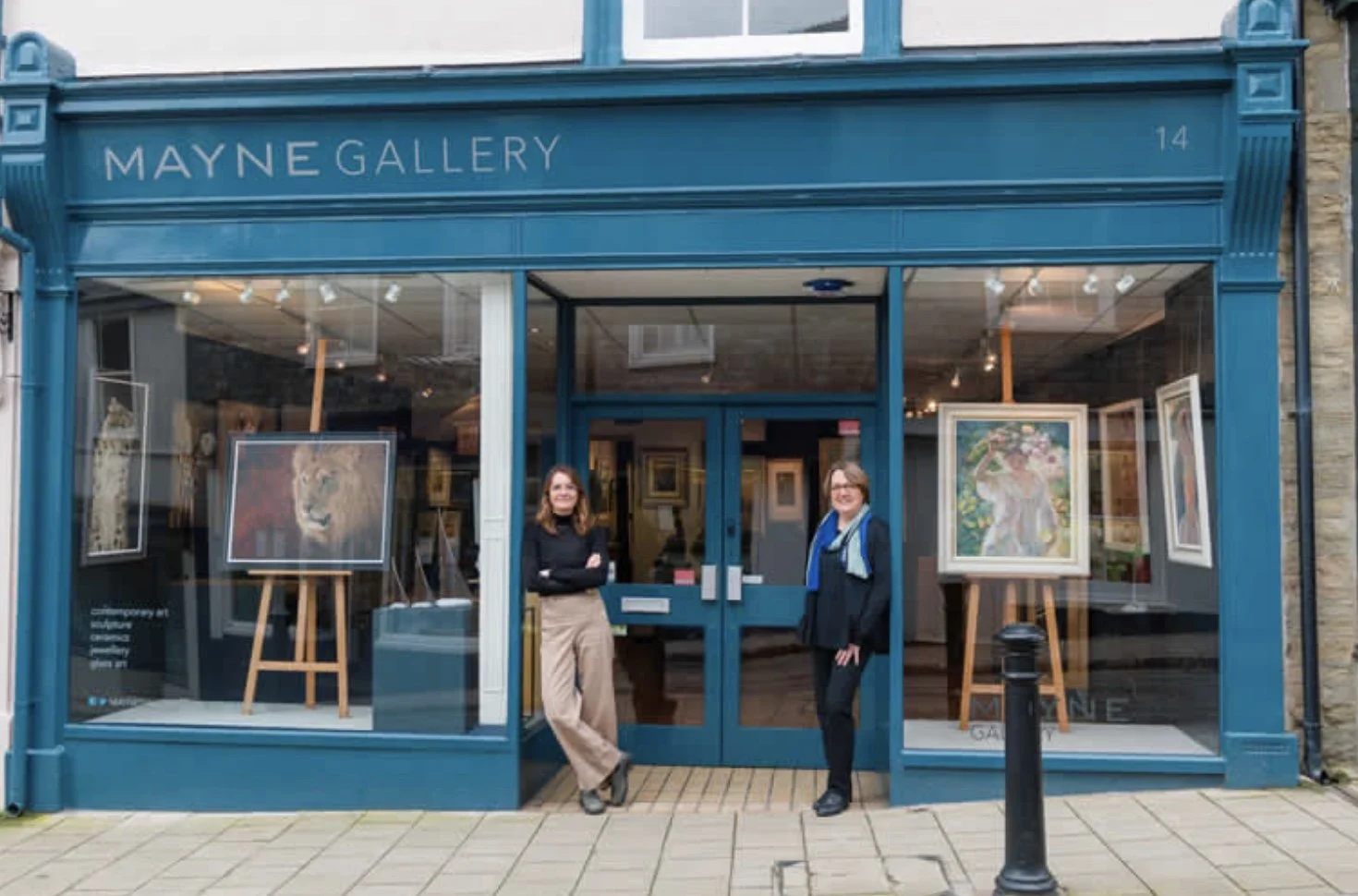 Two women standing in front of the Mayne Gallery art shop with paintings displayed in the window., and sculptures by Jonathan Hateley.