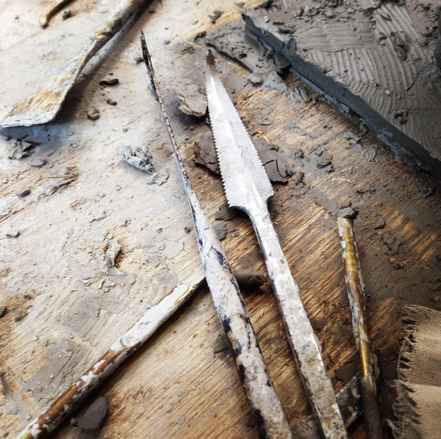 Close-up of artist's sculpting tools, including a metal loop tool, a serrated metal tool, and a stiff metal knife, resting on a wooden work surface covered with wet clay and clay scraps.