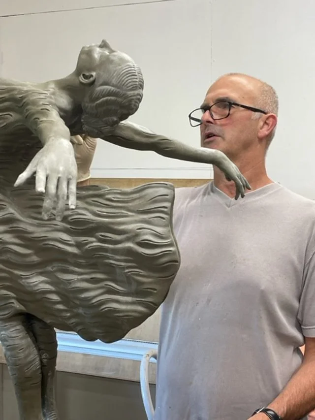 A man with glasses and a light gray shirt looking at a sculpture of a baby elephant sitting on a large leaf.