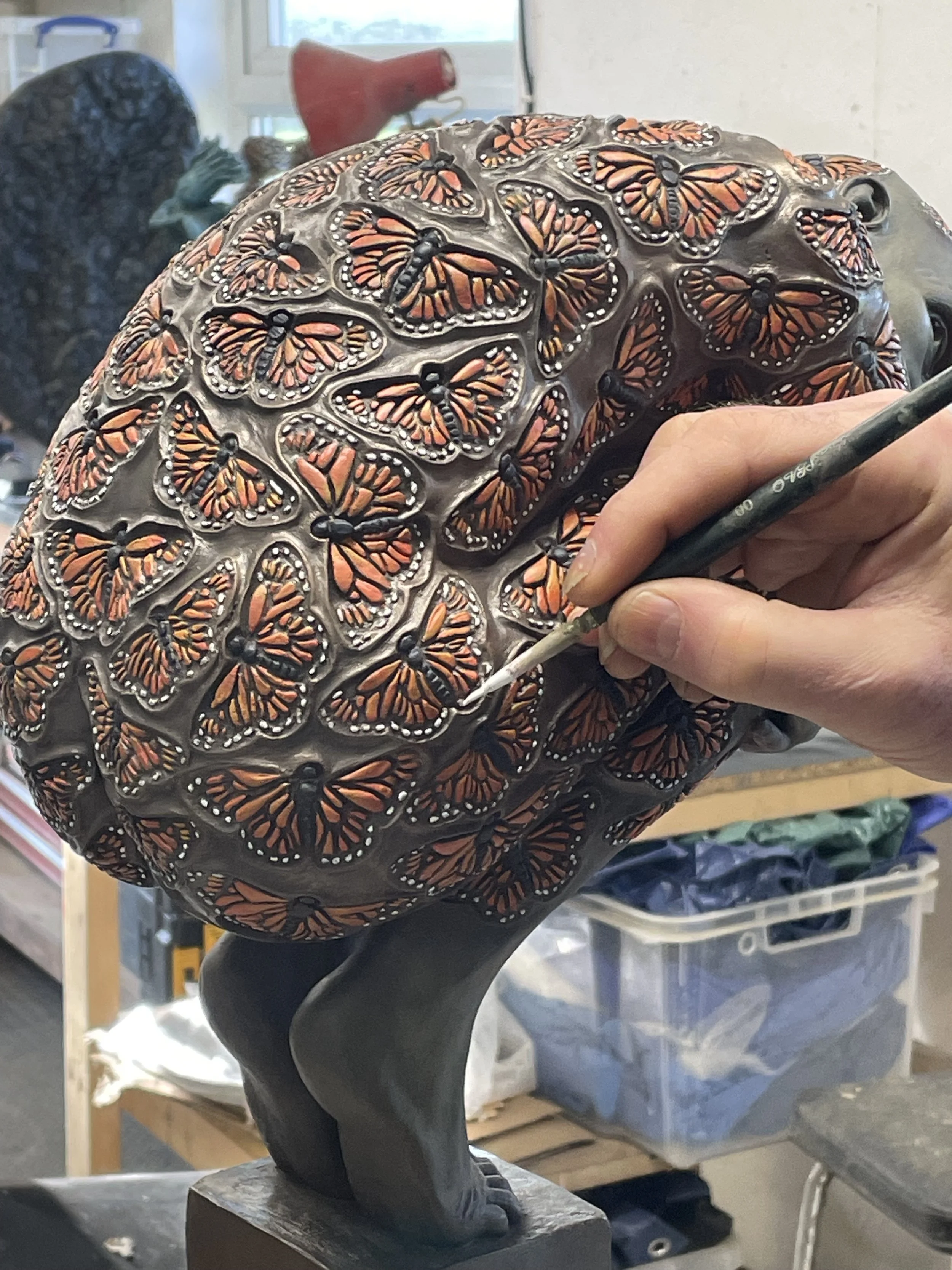 A sculptor working on a dark-colored sculpture of an animal skull, painting numerous butterflies with orange and black wings on the surface using a fine brush.
