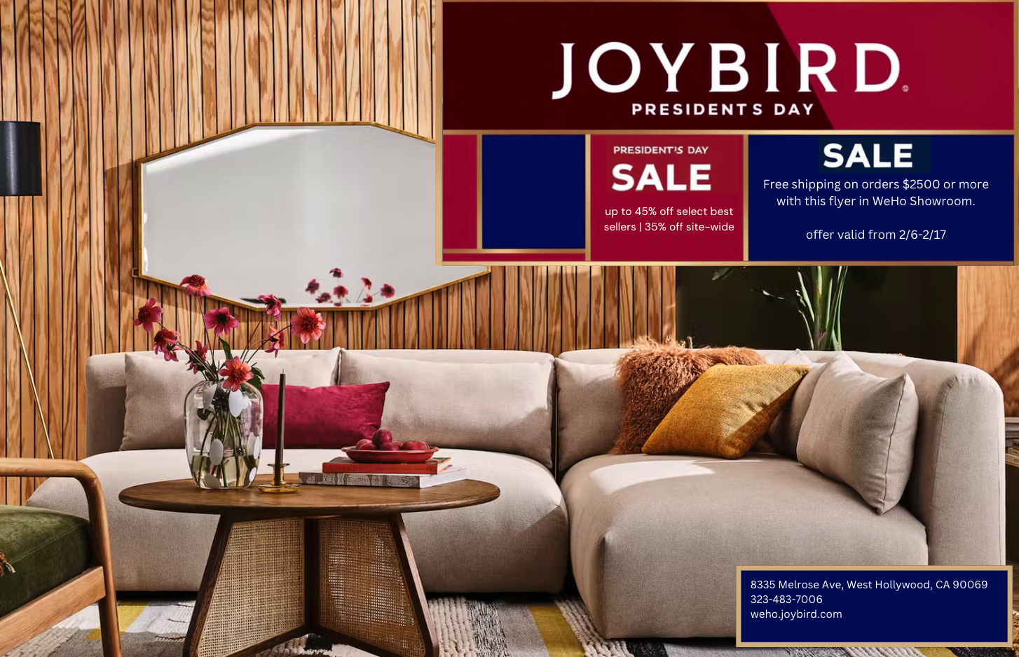 Living room with a beige sectional sofa, a wooden coffee table with a glass vase of pink flowers and red apples, and a rectangular mirror on wooden panel wall. A black floor lamp is on the left. A flyer for Joybird's Presidents Day sale on top right,
