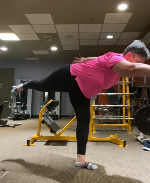 A woman in a pink shirt and black leggings performs a balance exercise on one foot in a gym, extending her arms forward while standing on a yellow exercise balance board.