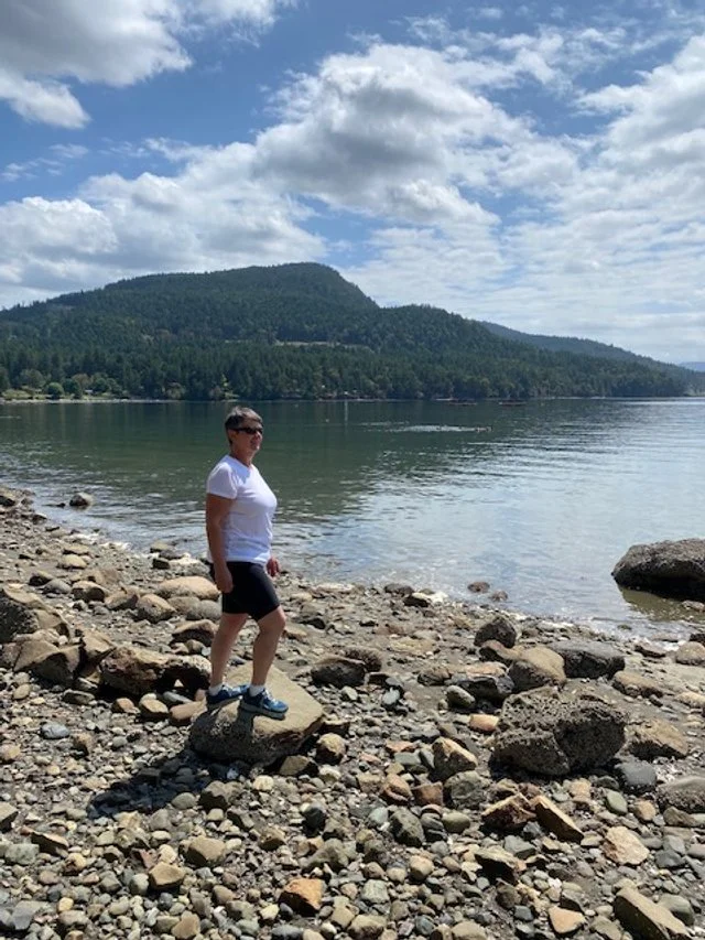 A woman standing on rocks by a lake with a mountain and forest in the background under a partly cloudy sky.