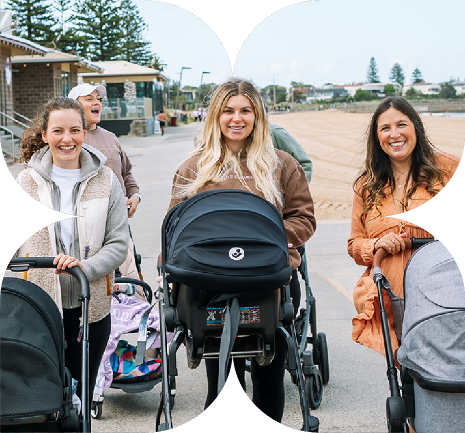 Four smiling women walking outdoors with strollers, in what appears to be a park or recreational area, on a cloudy day.
