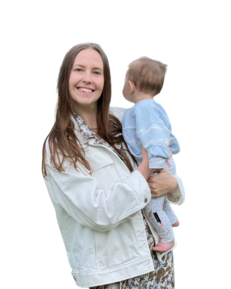 A woman with long brown hair holding a young child with short blond hair, smiling at the camera. The woman is wearing a white jacket and patterned dress, and the child is wearing a light blue shirt and patterned pants.