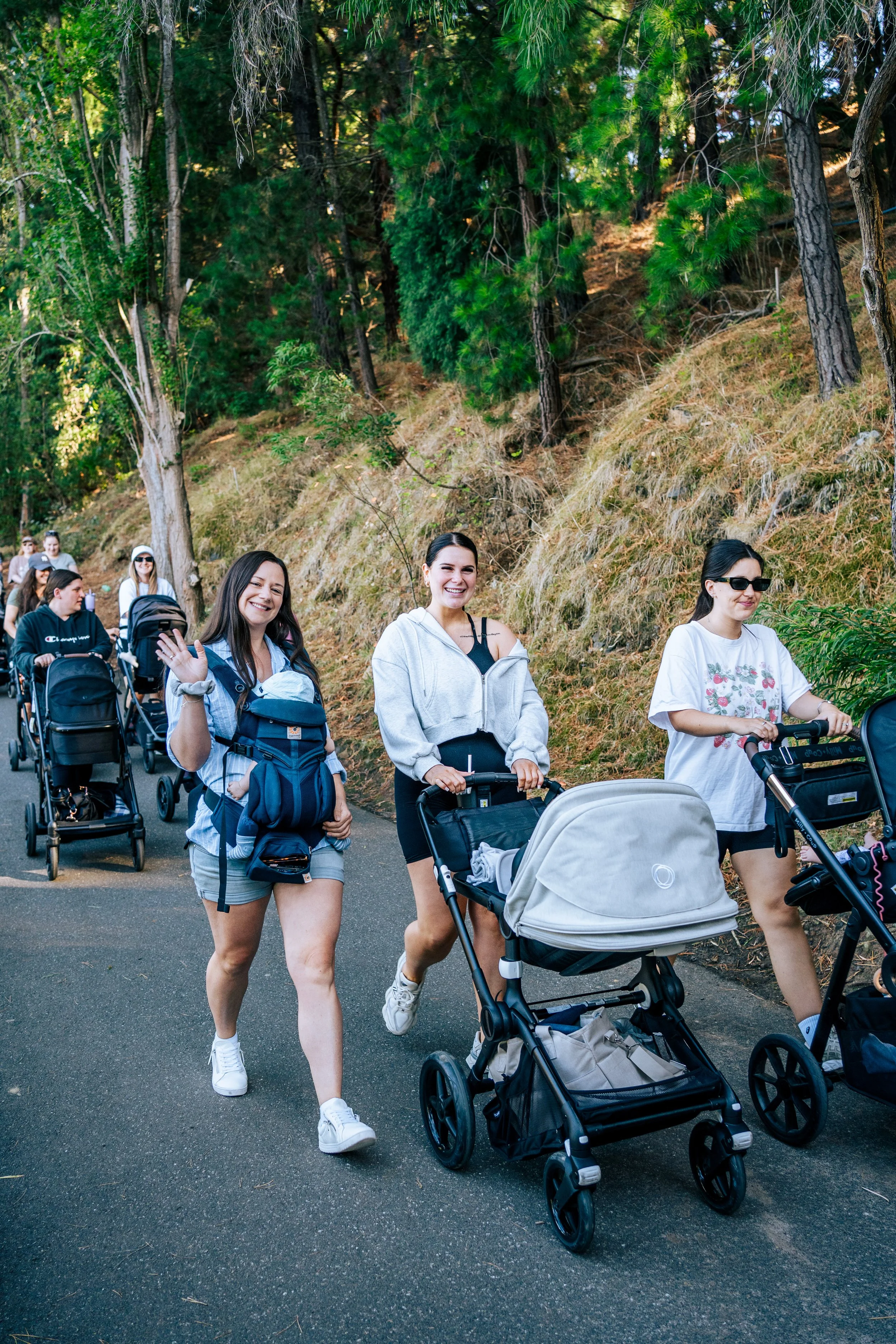 Smiling women walking with strollers and babies along a wooded path.