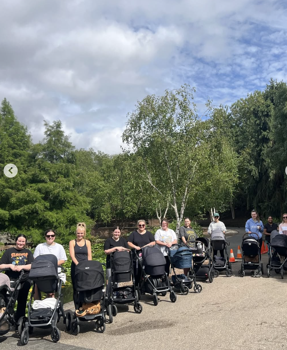 A group of women with strollers standing outdoors on a paved area, with trees and a blue sky with clouds in the background.